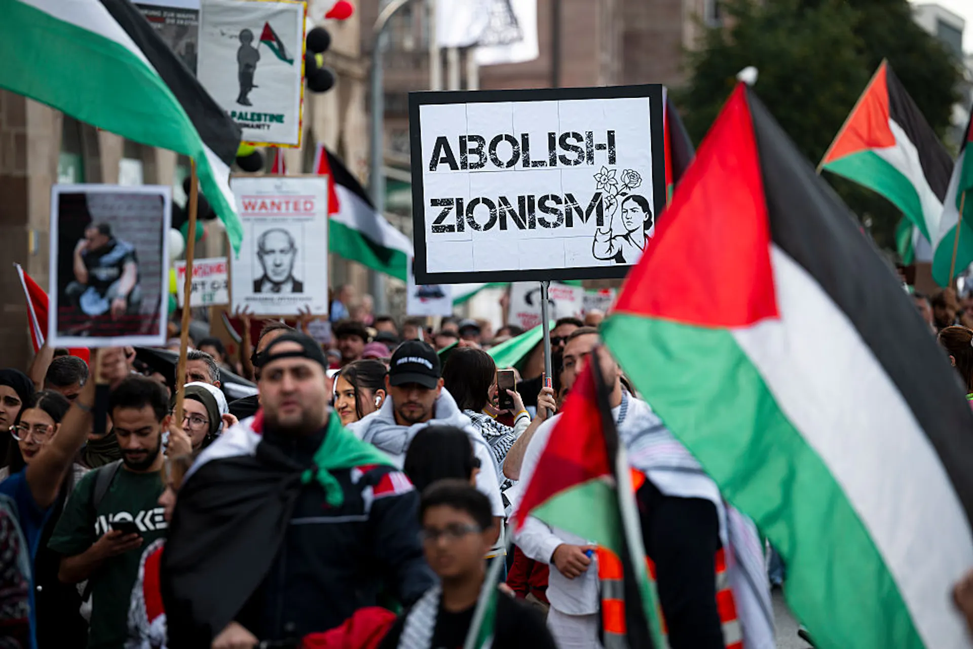 A street full of people marching with Palestinian flags and a sign that says 'Abolish Zionism.'